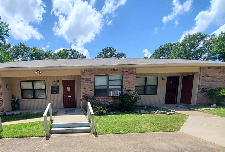 Single-story building with a brick facade, featuring multiple doors and windows. Concrete pathways lead to small stairways with railings at each entrance. Set in Pearl, Mississippi, this affordable housing complex boasts a backdrop of trees and a blue sky dotted with clouds.