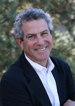 A person with curly gray hair, wearing a white shirt and dark blazer, smiling outdoors with greenery in the background.