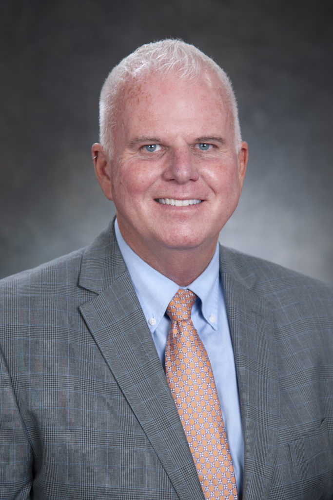 A man with short white hair, wearing a light blue shirt, orange patterned tie, and gray plaid suit jacket, smiling with an air of leadership in front of a gray background.