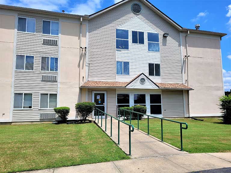 A three-story beige building with several windows and a glass entrance door, featuring a ramp for accessibility. This affordable housing in Jackson, Mississippi, is surrounded by a well-maintained lawn and shrubbery. The sky is clear and blue.