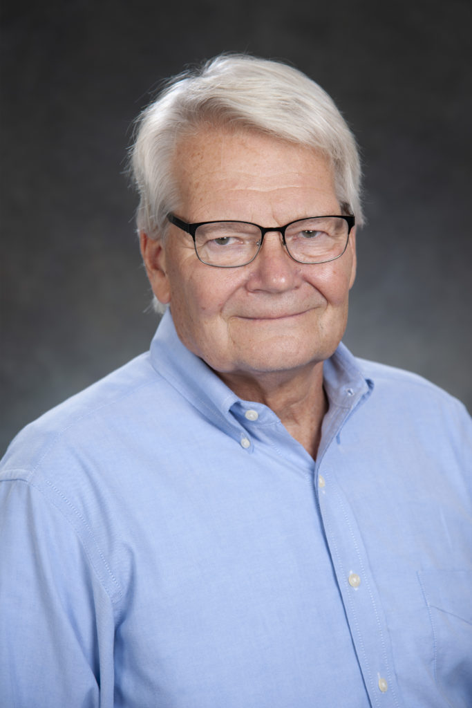 A man with white hair and glasses, exuding an air of leadership, is wearing a light blue button-up shirt and poses confidently against a dark gray background.