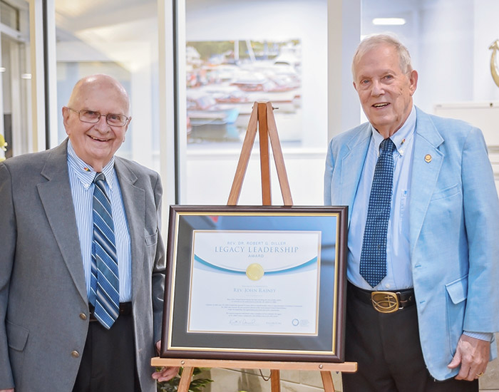 Two elderly men dressed in suits stand on either side of a framed Legacy Leadership Award displayed on an easel. Indoor setting with a blurred painting in the background.