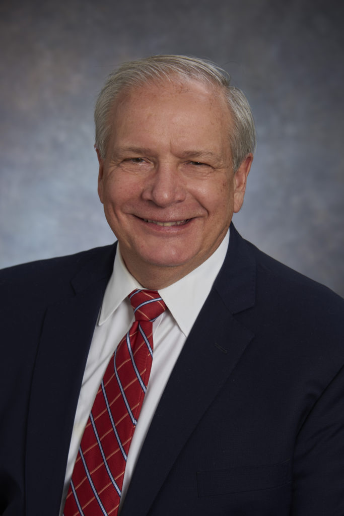 A man dressed in a dark suit with a white shirt and red striped tie, exuding leadership as he smiles at the camera against a neutral background.