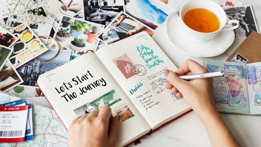 A person is writing travel plans in a journal with a cup of tea nearby. Surrounding the journal are photos, maps, postcards, and travel tickets. The journal entry starts with "Let's Start The Journey.