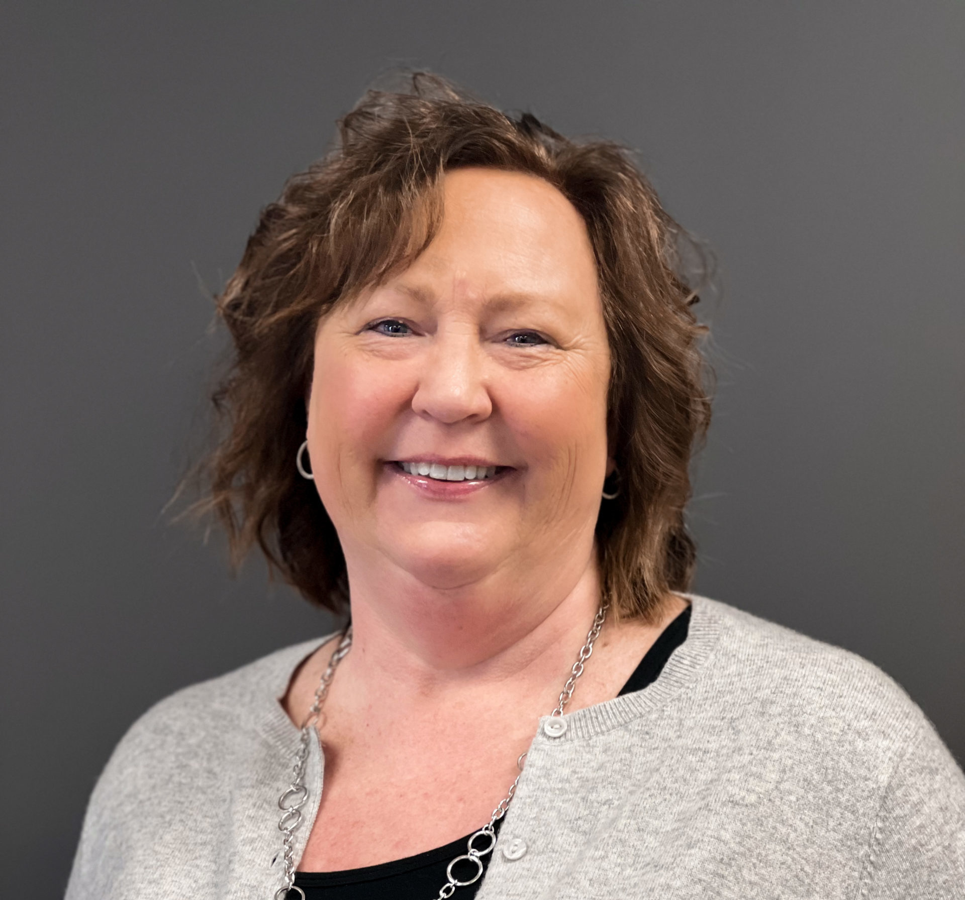 A woman with shoulder-length brown hair smiles while wearing a light gray cardigan and a black top against a plain dark background, as The Polaris Community welcomes new leadership.