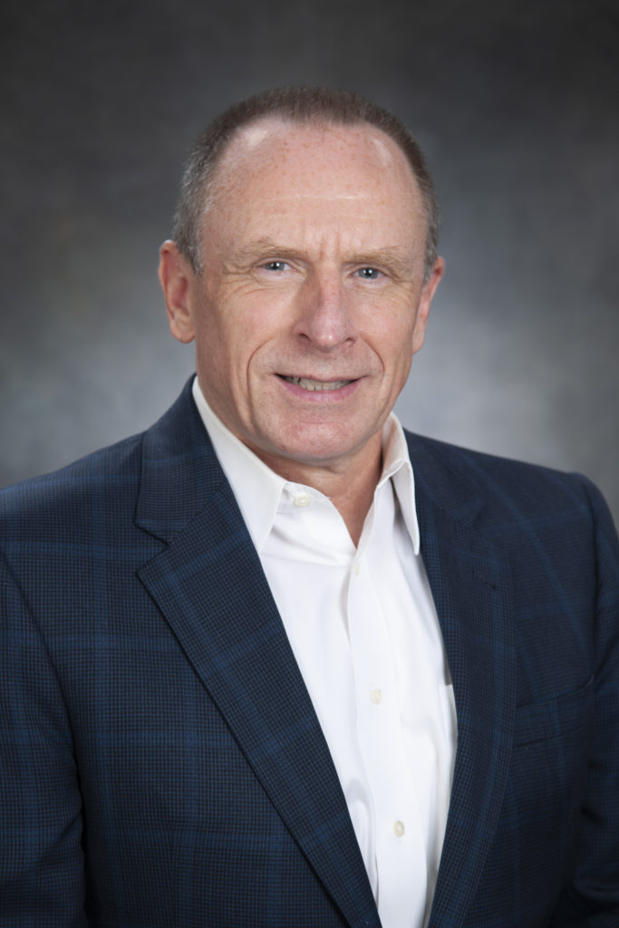 A close-up of a middle-aged man with short hair wearing a dark suit jacket and a white dress shirt, smiling confidently, embodying leadership. Neutral background.