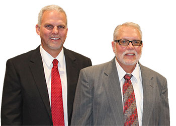 Two men in suits standing together; one in a black suit with a red tie and the other in a gray suit with a patterned red tie. Both have gray hair and glasses, and are smiling at the camera.