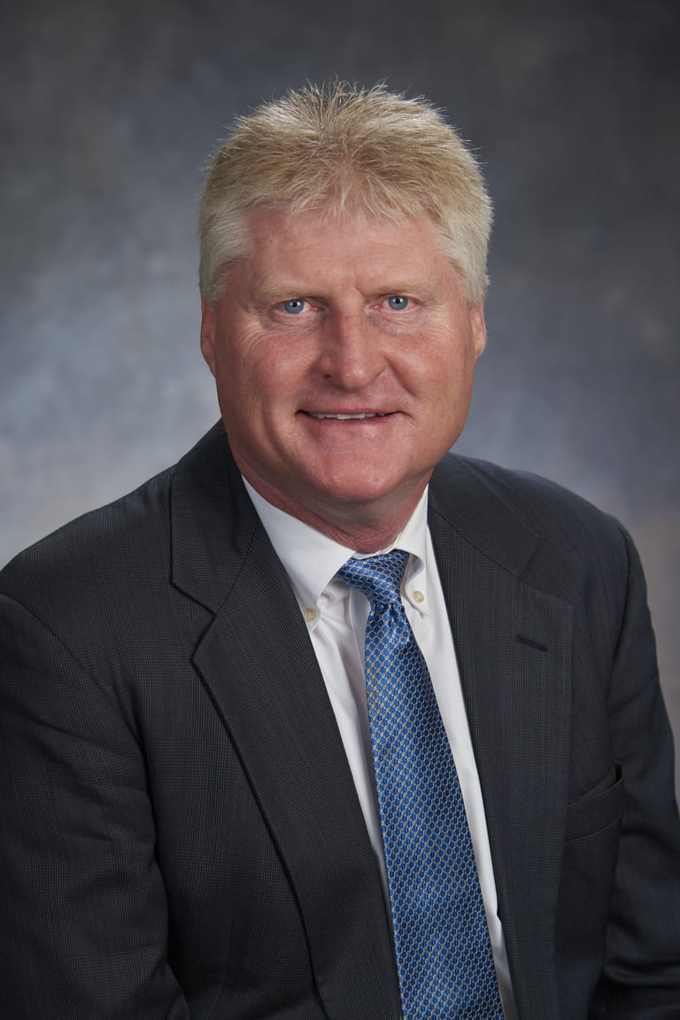 A man with short blond hair in a dark suit, white dress shirt, and blue tie, exuding leadership as he smiles confidently at the camera against a gray studio background.