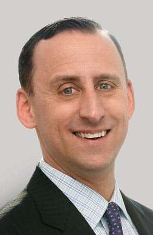 A man with short dark hair, exuding leadership in his suit, shirt, and tie, smiles confidently at the camera against a plain light background.