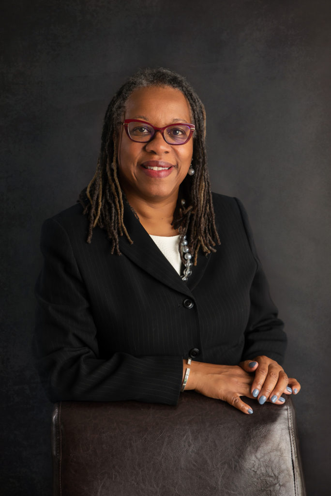 A woman with glasses and shoulder-length dreadlocks, exuding leadership in a black blazer, sits with her arms resting on the back of a chair against a dark background.