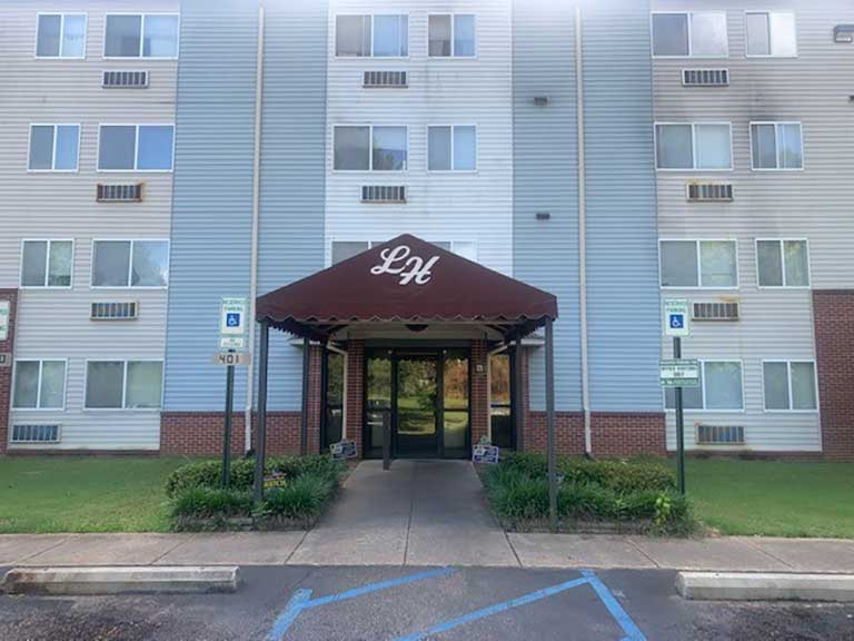 Front entrance of a multi-story residential building with an awning displaying the letters "LH." Accessible parking spaces and a sidewalk leading to the entrance are visible. This represents part of the affordable housing in Jackson, Mississippi.