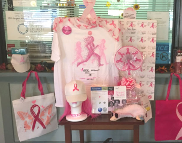 A display features various breast cancer awareness items including a T-shirt, cap, tote bag, brochures, and a small stuffed animal, all adorned with pink ribbons and other related designs.