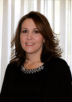 A woman with shoulder-length brown hair is standing in front of a window with white curtains, wearing a black top with a beaded neckline.