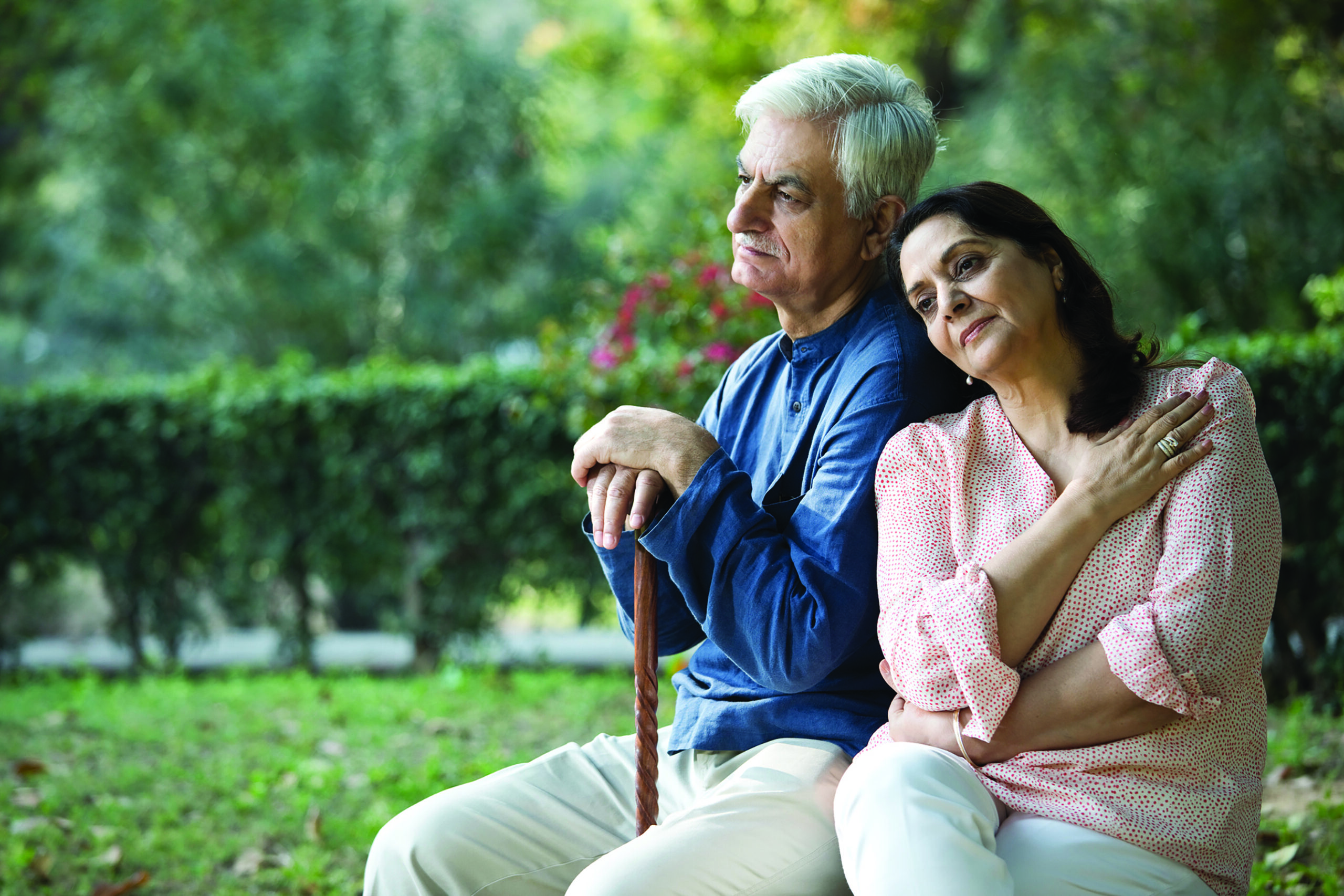 An elderly couple sits on a bench in the park, the woman leaning her head on the man's shoulder as he gently holds his cane, savoring a moment of togetherness amidst life's bittersweet loneliness.