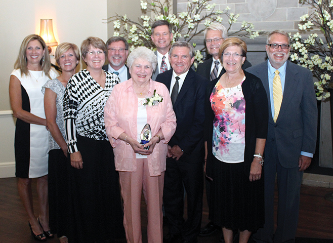 A group of ten formally dressed adults posing indoors, with a white-haired woman in the center holding an award.