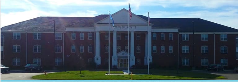 A three-story brick building labeled "Evans Hall II" with tall white columns at the entrance, displaying three flags in front on a sunny day, serves as an emblematic structure of affordable housing in Dayton, Ohio.