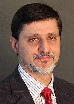 A middle-aged man with short dark hair and a neatly trimmed beard is wearing a suit and a red tie, posing against a plain dark background.