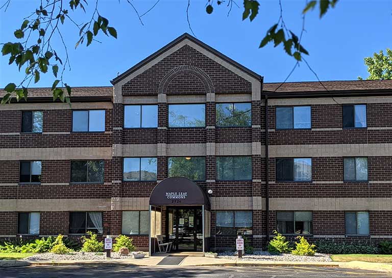 A three-story brick building with a dark-colored arched entrance stands proudly as "Mainstreet Commons." This charming residence, offering affordable housing in Goshen, Indiana, features small trees and plants that add character and warmth to its surroundings.