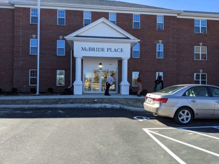 A three-story brick building labeled "McBRIDE PLACE" with a white entrance portico, several people at the entrance, and a parked car in the foreground showcases affordable housing in Dayton, Ohio.