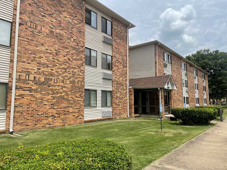 A three-story brick and siding apartment building with windows and a small entrance canopy, surrounded by grass and shrubs under a cloudy sky, embodies the essence of affordable housing in Clarksdale, Mississippi.