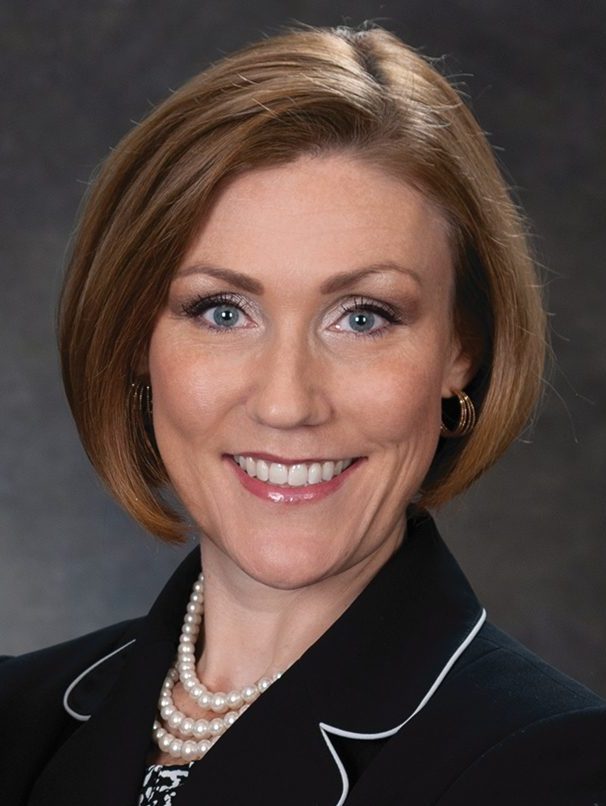 A woman with short brown hair smiles confidently, wearing a dark blazer that exudes leadership, complemented by a pearl necklace and earrings, set against a dark background.