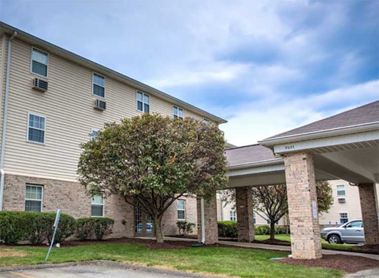 A four-story cream-colored apartment building with a covered carport area and greenery, including a large tree and shrubs, stands prominently under a cloudy sky, representing affordable housing in Uniontown, Pennsylvania.