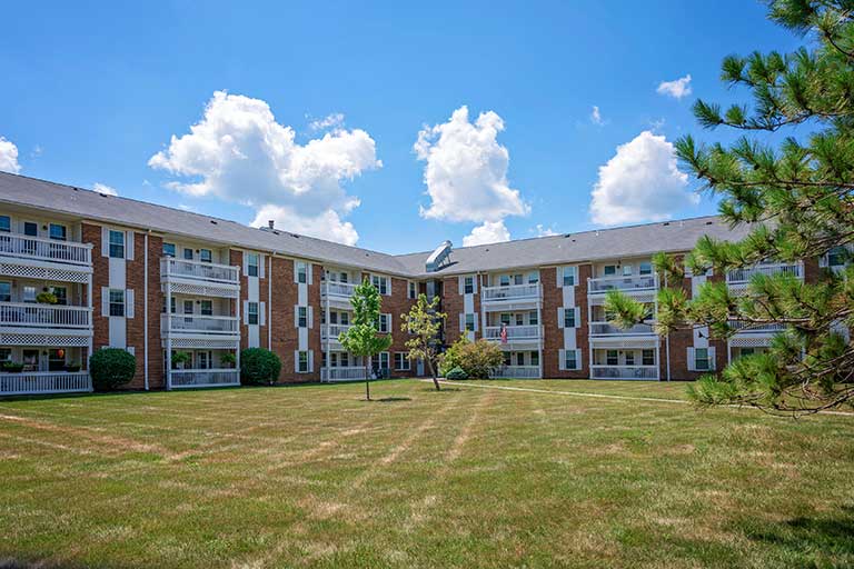 A three-story brick apartment building forms a U-shape around a grassy courtyard with a few small trees under a partly cloudy sky.