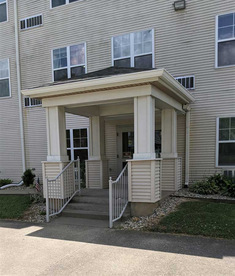 An affordable housing option in New Albany, Indiana, this beige apartment building features a small covered entrance with stairs and railings. The building also has windows and charming landscaping with rocks and plants around the entrance.