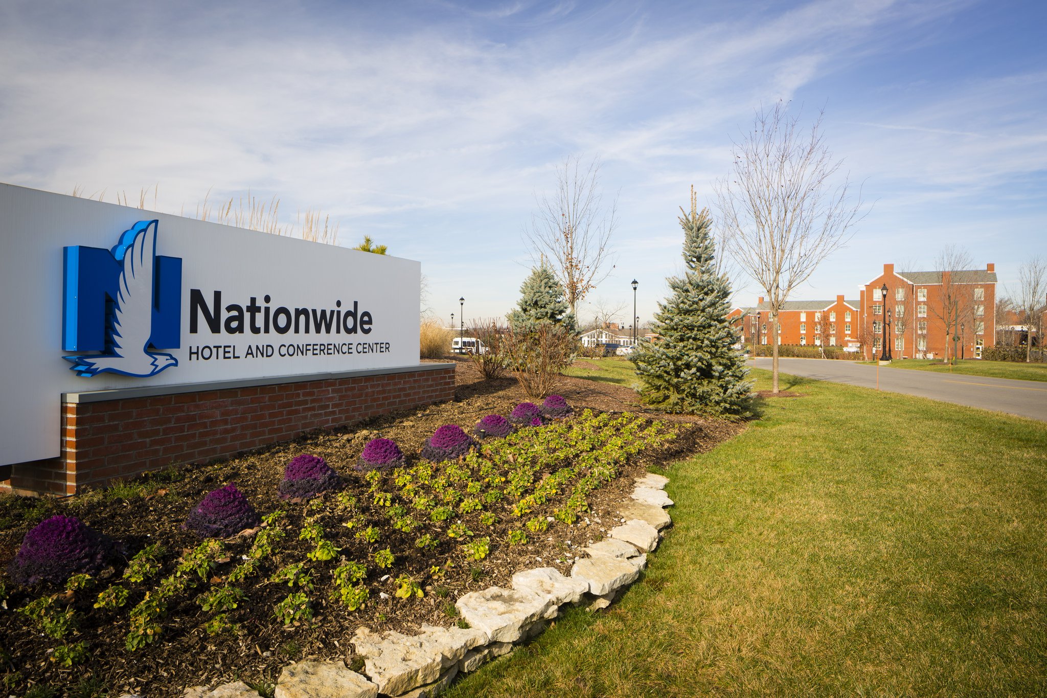 Sign for Nationwide Hotel and Conference Center next to landscaped garden with purple flowers and evergreen trees, with a brick building visible in the distance under a partly cloudy sky.