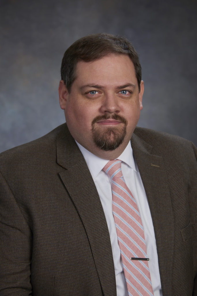 A man with short hair, a beard, and mustache is wearing a brown checked suit jacket, white shirt, and pink striped tie, exuding leadership as he poses for a professional portrait against a dark background.