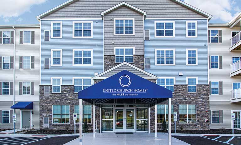 Four-story building with gray and blue siding, highlighted by a blue awning at the entrance displaying "United Church Homes" under a logo.