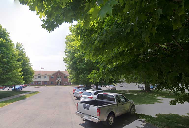 A parking lot with several vehicles is shaded by overhanging tree branches. Nearby, a three-story red brick building with a small steeple stands out, highlighting the charm of affordable housing in Bremen, Indiana.