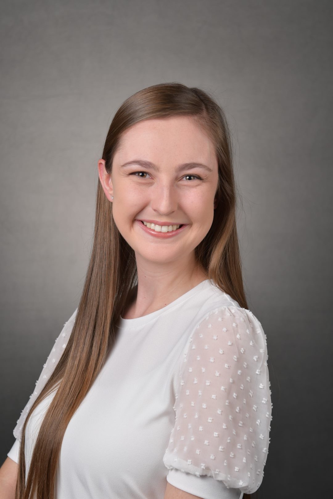 A young woman with long, straight hair wearing a white blouse smiles confidently, representing the fundraising team in front of a grey background.