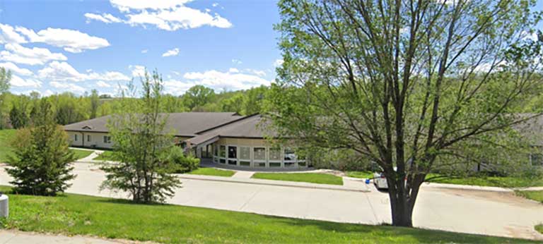 A one-story building with beige exterior walls surrounded by trees and greenery under a partly cloudy sky. A driveway and a few small pine trees are in the foreground, highlighting the charm of affordable housing in Macy, Nebraska.