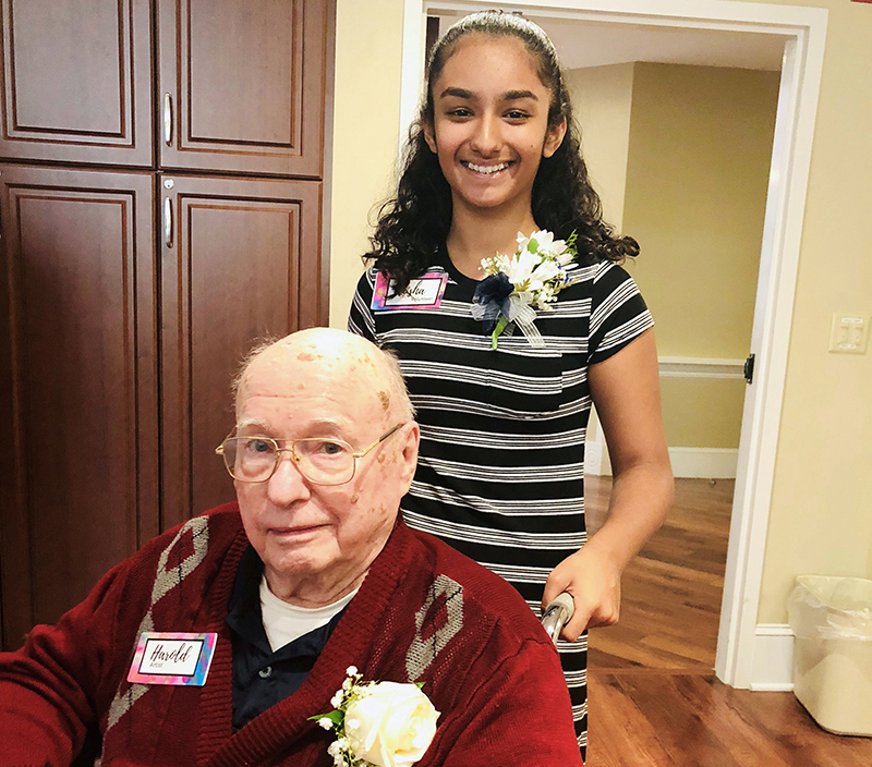 An elderly man sits in a wheelchair wearing a red sweater and glasses, with a young girl standing behind him. Both are smiling and wearing name tags and floral boutonnières.