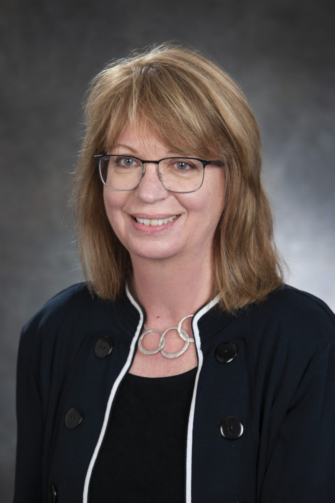 A woman with shoulder-length light brown hair, wearing glasses, a black top, and a dark jacket with white trim, smiles confidently in front of a gray background, embodying leadership.