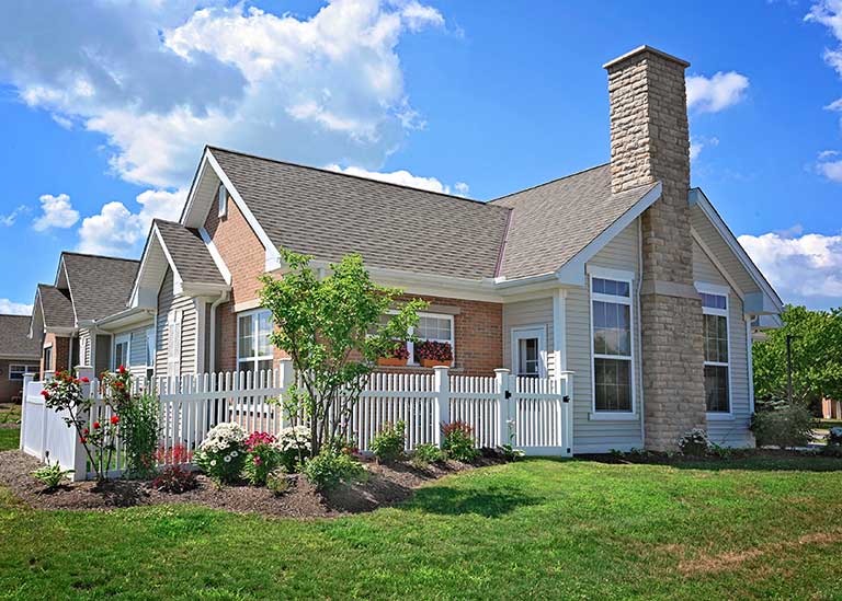 Single-story house with a gabled roof, brick and siding exterior, a stone chimney, and a white picket fence surrounding a small garden with flowers. Background shows a grassy lawn and a partly cloudy sky.