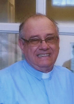 An elderly man wearing glasses and a light blue clerical shirt with a white clerical collar. He is standing in front of a building's window or door.