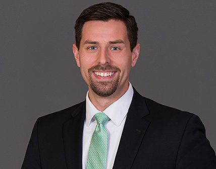 A man with short dark hair and a trimmed beard, wearing a dark suit, white shirt, and green tie, smiles at the camera against a gray background. His confident demeanor reflects his expertise on topics like the SECURE Act.