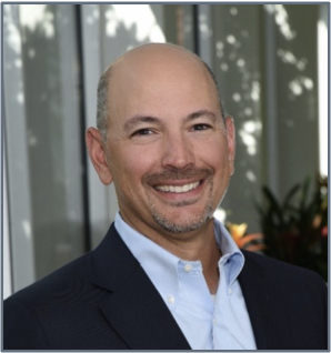 A person with a bald head and light beard is smiling, exuding leadership while wearing a dark jacket and light blue shirt. They stand confidently in front of a building with glass windows and greenery in the background.