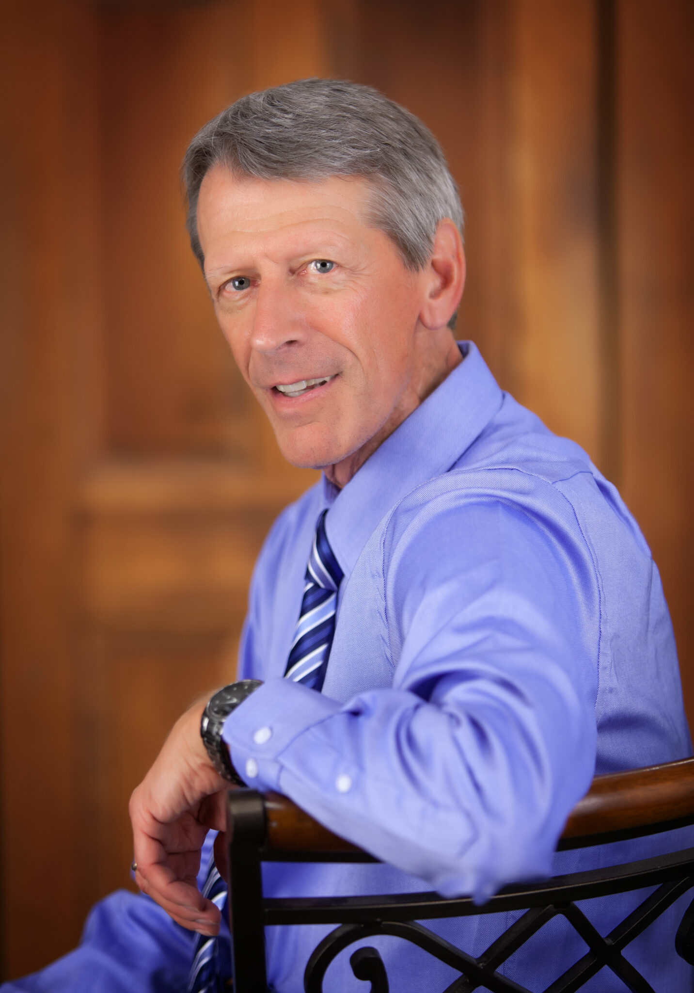 A man with short gray hair wearing a blue dress shirt and striped tie sits on a chair, looking at the camera with a slight smile, embodying the professionalism of our dedicated fundraising team.