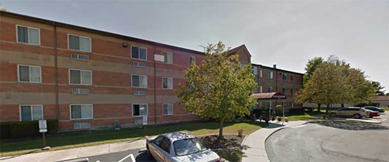 A three-story brick building with many windows, a curved driveway, and a few parked cars. Trees are situated in front of the building, offering an inviting look to this affordable housing in Pickerington, Ohio.
