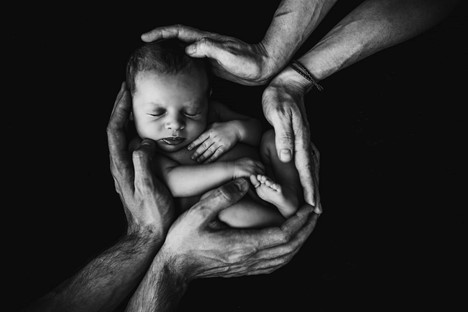 A newborn baby is cradled by multiple adult hands in a protective and nurturing gesture, set against a dark background.