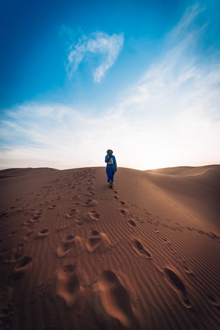 A person dressed in blue walks up a sand dune, leaving footprints behind under a clear blue sky with some clouds.