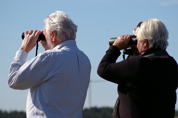 Two elderly individuals, one man and one woman, stand outdoors looking through binoculars toward the horizon.