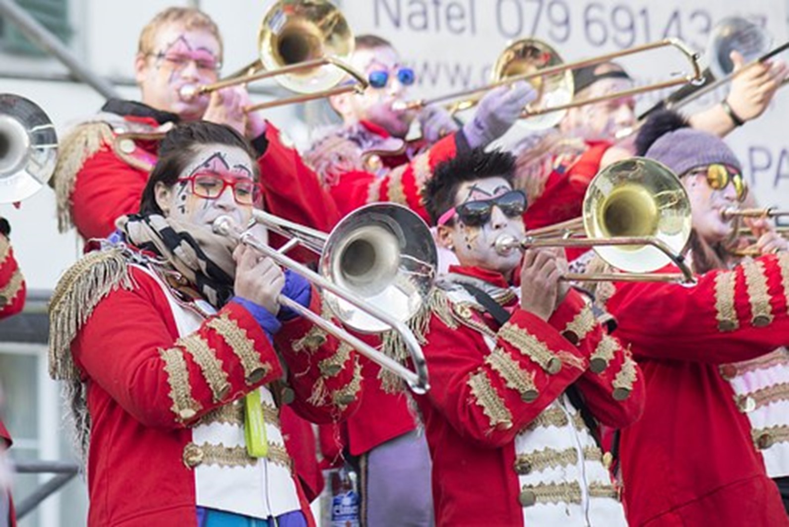 A group of musicians in bright red uniforms and face paint play trombones at an outdoor event.