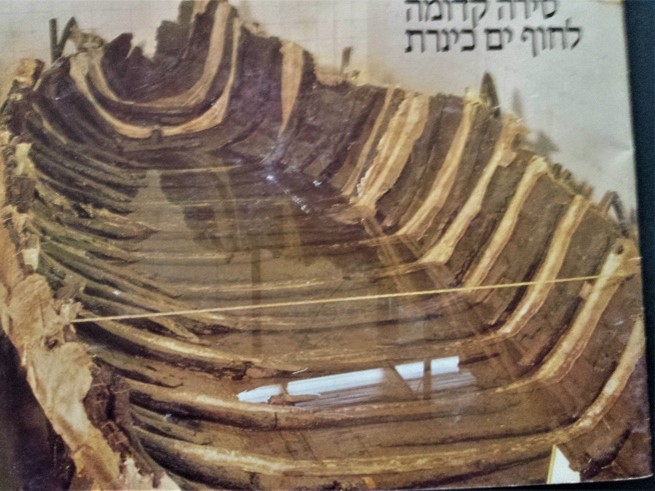 Photograph of an ancient wooden boat skeleton on display, with text in Hebrew visible in the upper right corner.