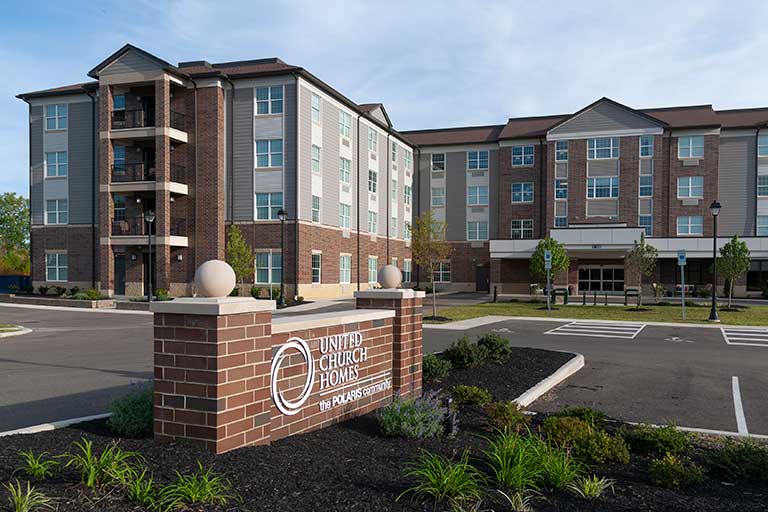 Image of a four-story brick building with a sign reading "United Church Homes The Polaris Community" in the foreground, featuring landscaped grounds and an adjacent parking lot.