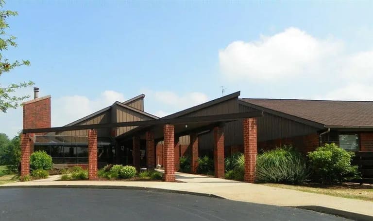 A single-story brick building with multiple sloping roofs and a covered walkway supported by brick columns, surrounded by greenery and a paved driveway, exemplifies the charm of affordable housing in Avilla, Indiana.