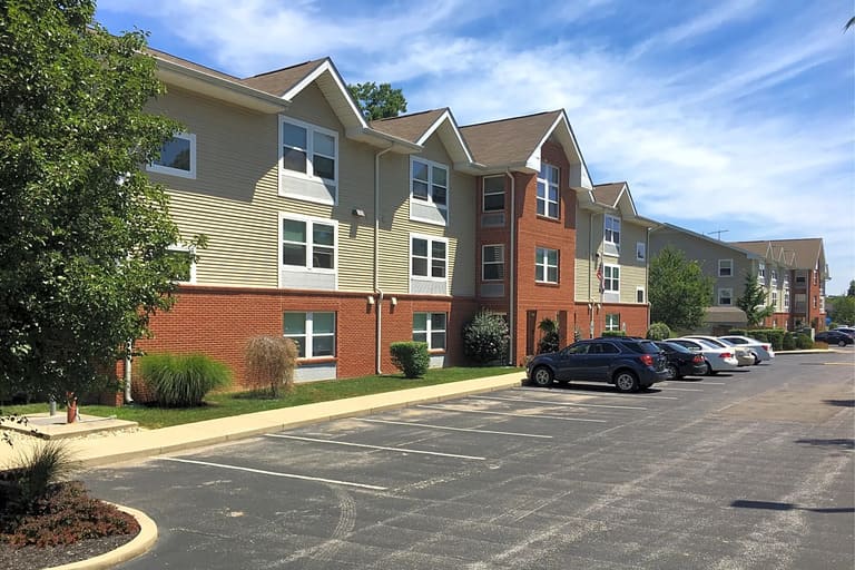 Three-story apartment building with beige and red brick exterior in affordable housing in Bloomington, Indiana. Several cars are parked in front, next to a sidewalk and lawn with bushes and trees. Clear blue sky overhead.